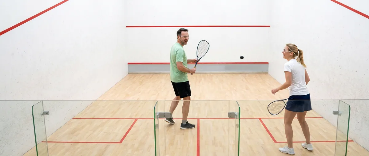 Twee spelers in een rally, gezien door de glazen achterwand van de squashbaan, de bal in de lucht